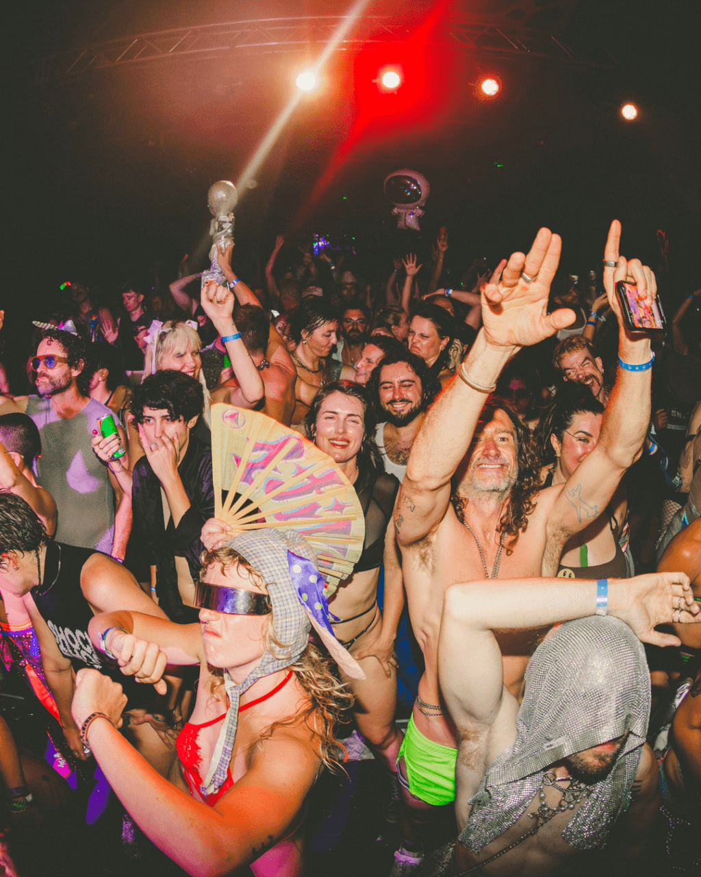 A large group of people dance energetically at a party, many wearing costumes, sunglasses, or headpieces under colorful stage lights, with hands raised and joyful expressions.