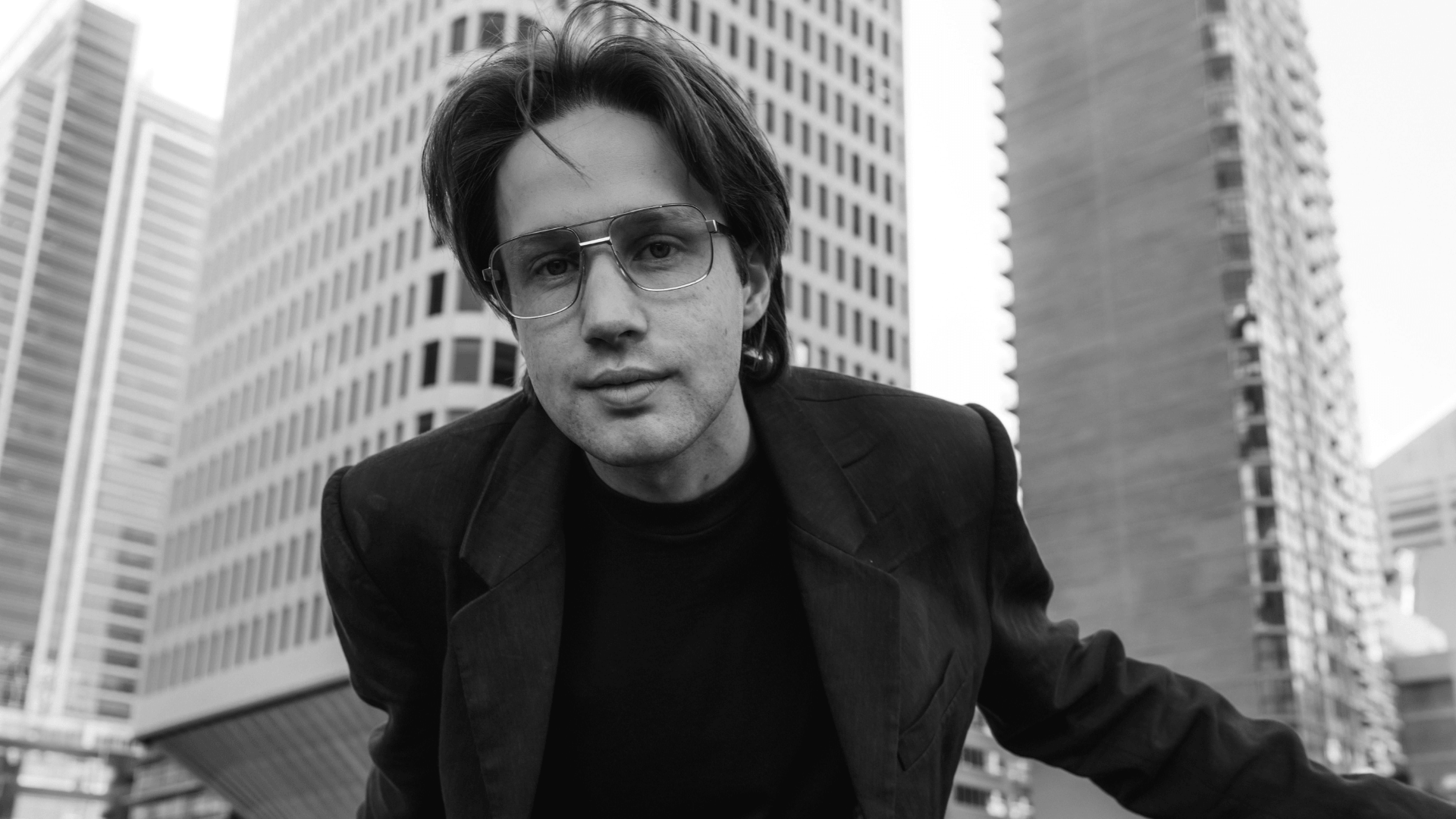 A person with medium-length hair and glasses, wearing a dark jacket, leans toward the camera in front of tall city buildings. The image is in black and white.