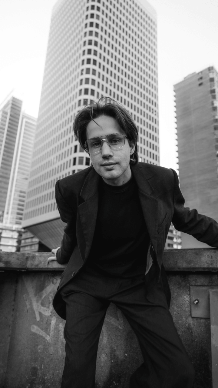 A person in a suit leans forward on a rooftop with tall office buildings in the background. The image is in black and white.