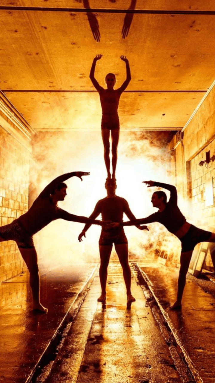 Four acrobats perform a balancing act in a tunnel, with one standing on anothers shoulders and two others posing beside them. Bright orange light and smoke create dramatic shadows on the wall behind them.