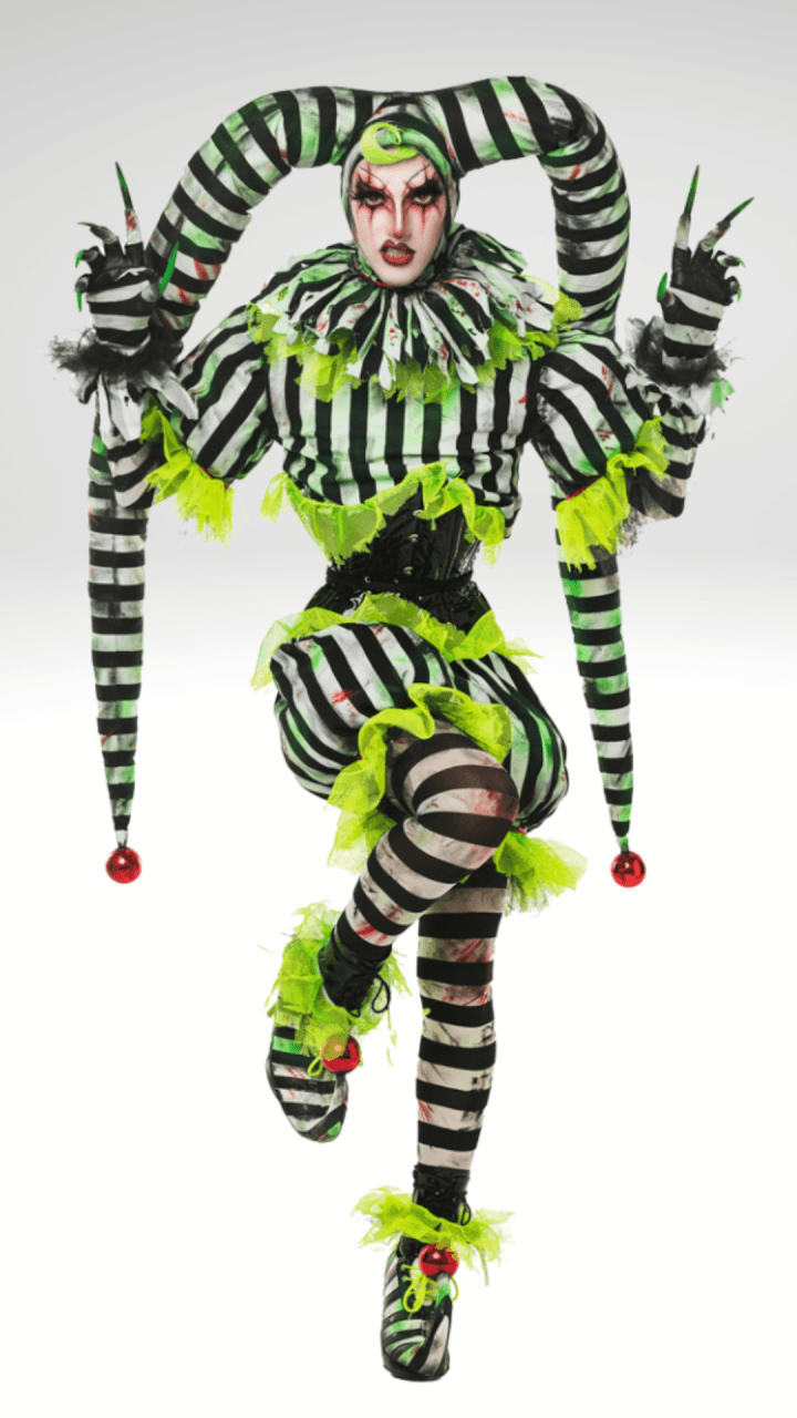 Person in a jester costume with black, white, and neon green stripes, pointed hat, ruffled collar, and dramatic makeup, standing on one leg with arms raised, against a plain background.