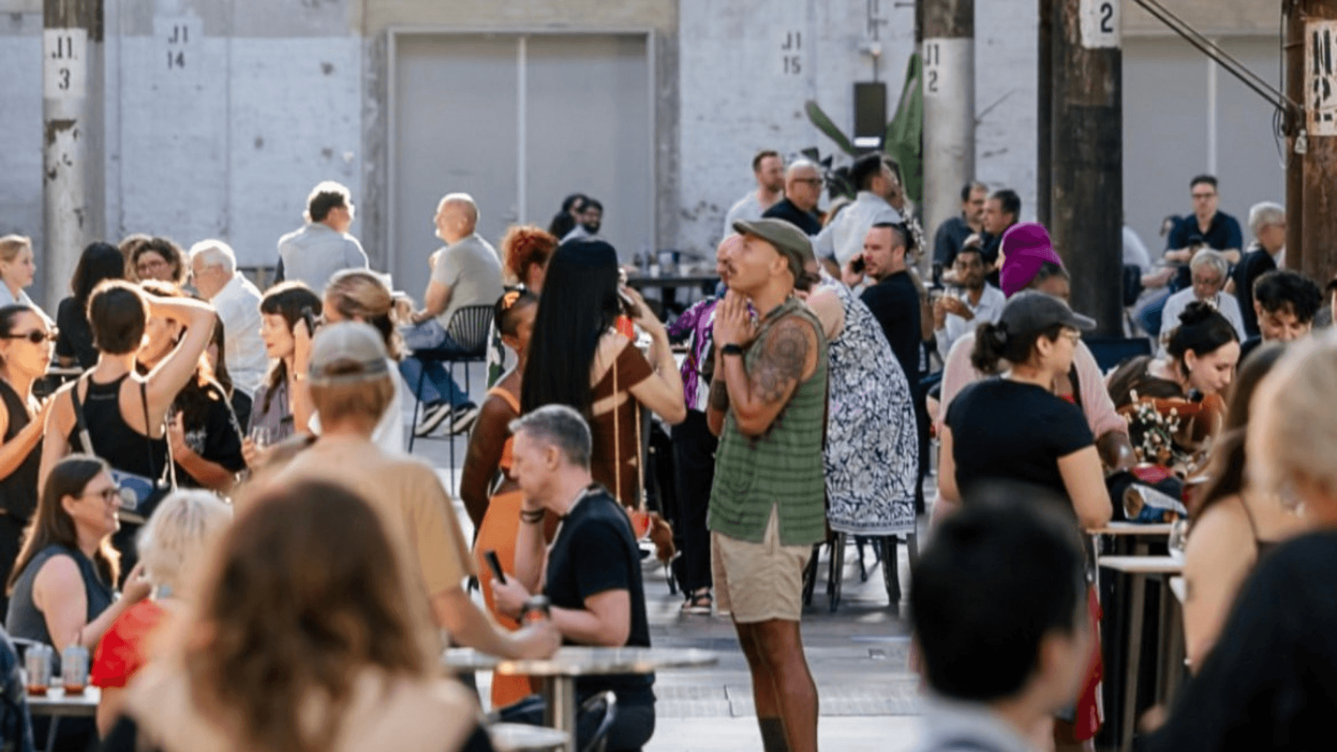 A busy outdoor cafe with many people sitting at tables and talking. One person stands in the center wearing a green shirt, shorts, and a cap. Sunlight brightens the lively scene.