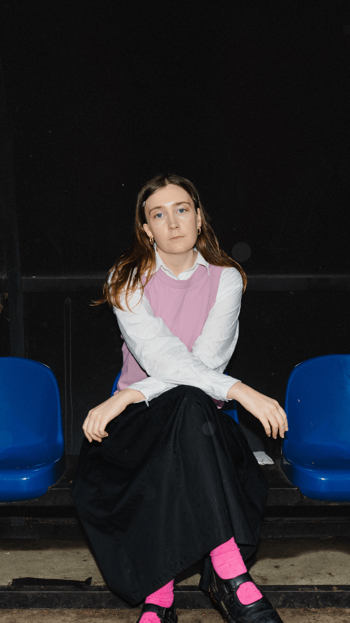 A woman with long brown hair sits on a blue chair, wearing a white shirt, pink vest, long black skirt, and bright pink socks with black shoes, against a dark background.