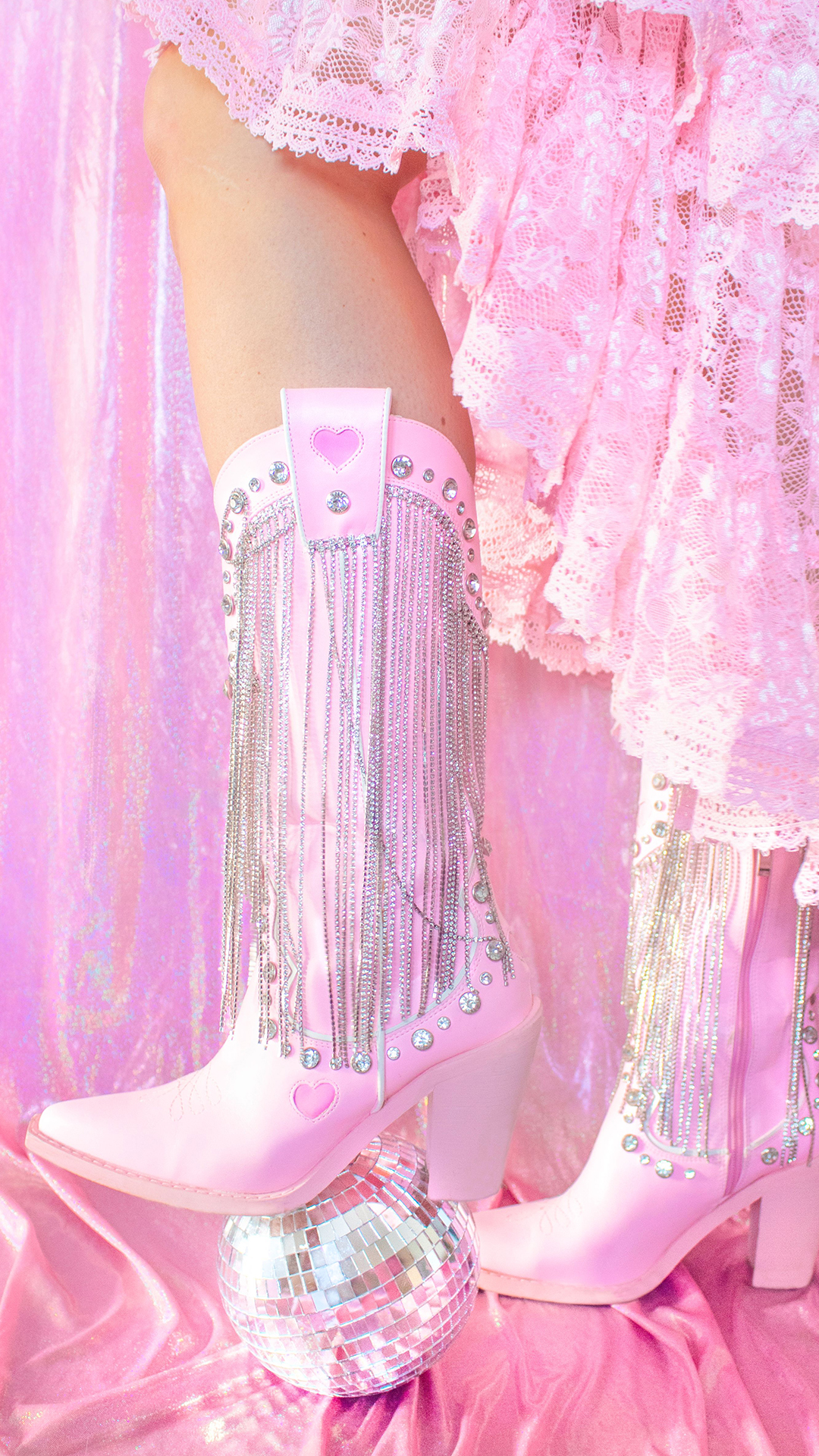 A person wearing pink cowboy boots with silver chains and a pink lace skirt stands with one foot on a small disco ball against a shiny pink backdrop.