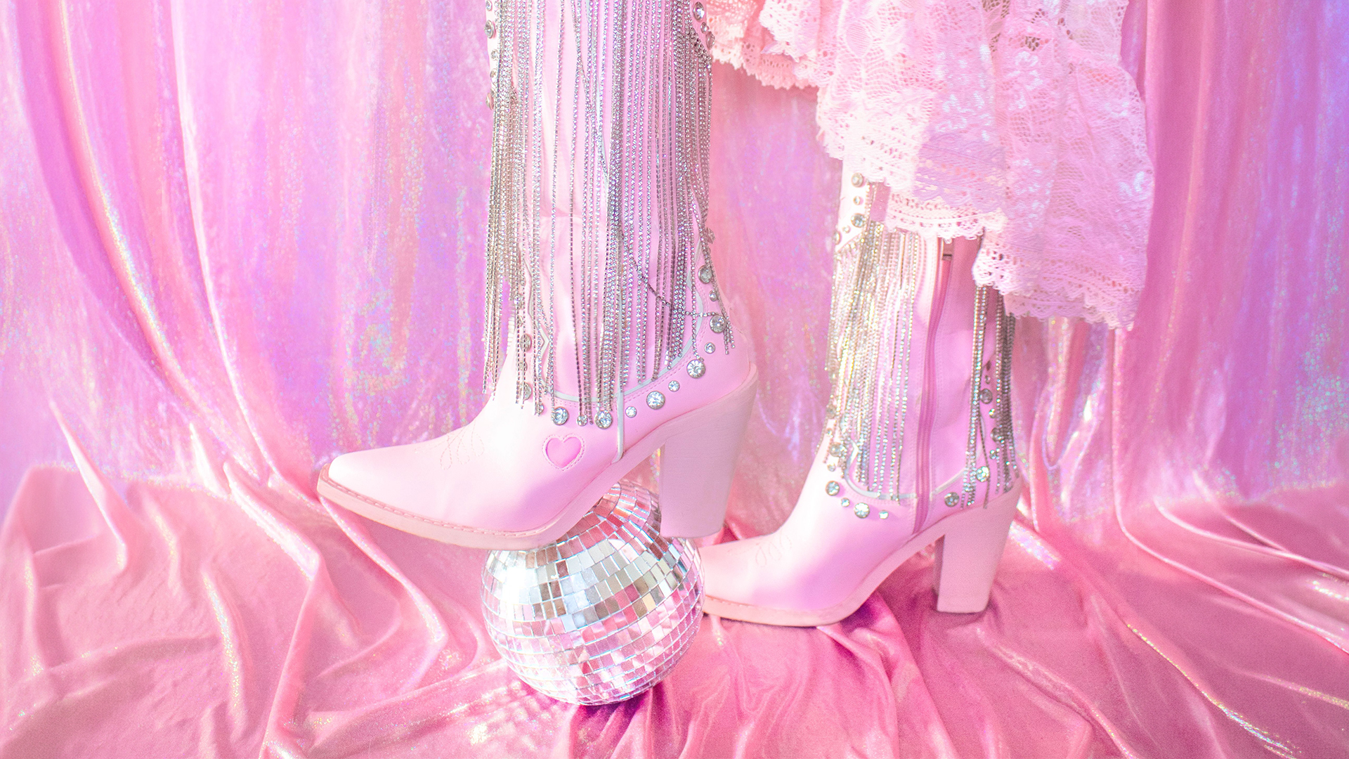 Person wearing pink heeled boots with silver chains and heart detail, standing on a disco ball. The background and clothing are shiny pink fabric and lace, creating a glamorous, playful scene.
