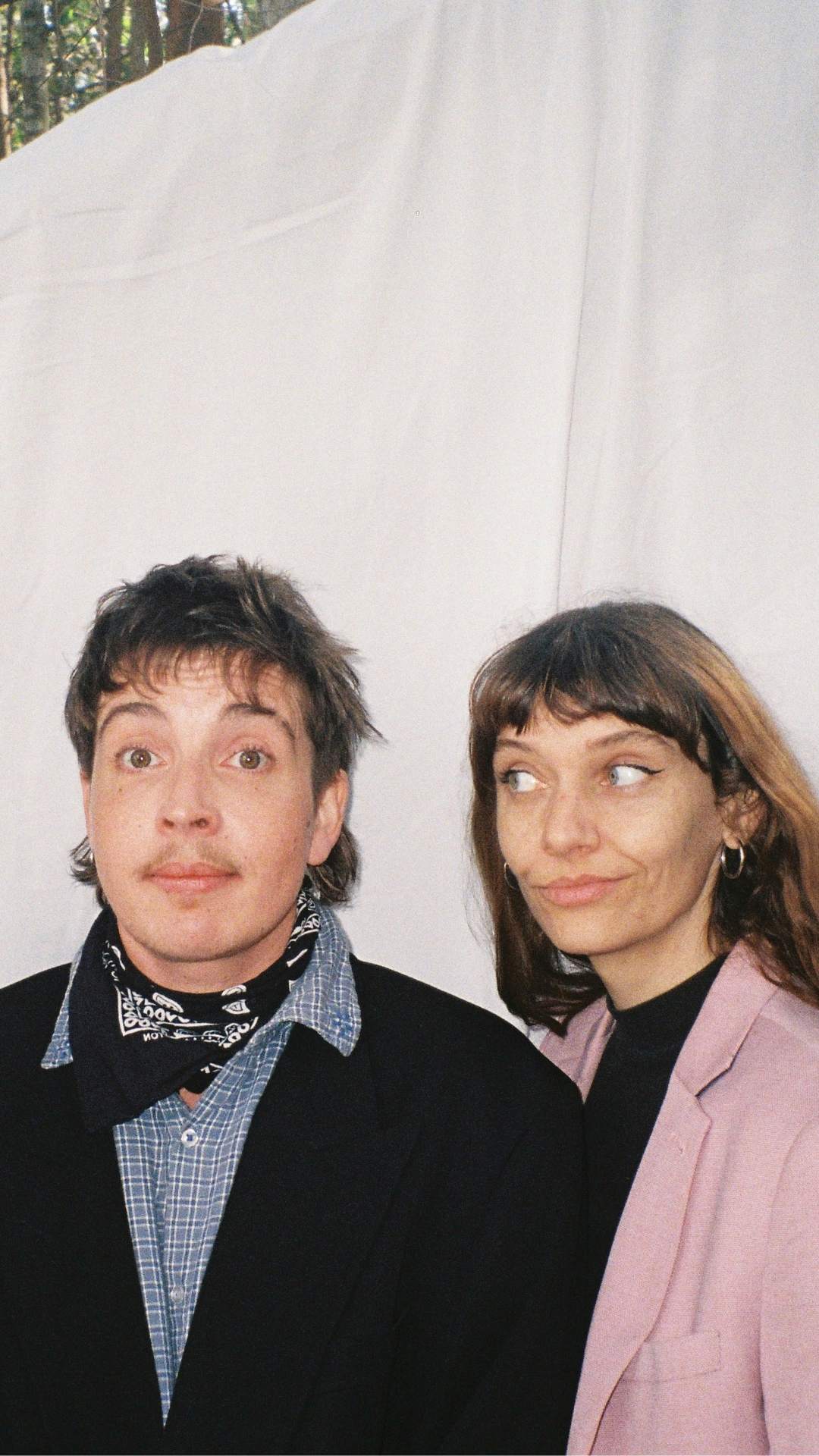 Two people stand side by side in front of a white sheet. One wears a black suit and bandana; the other wears a pink blazer. Both have amused expressions, with the person on the right looking sideways at the other.