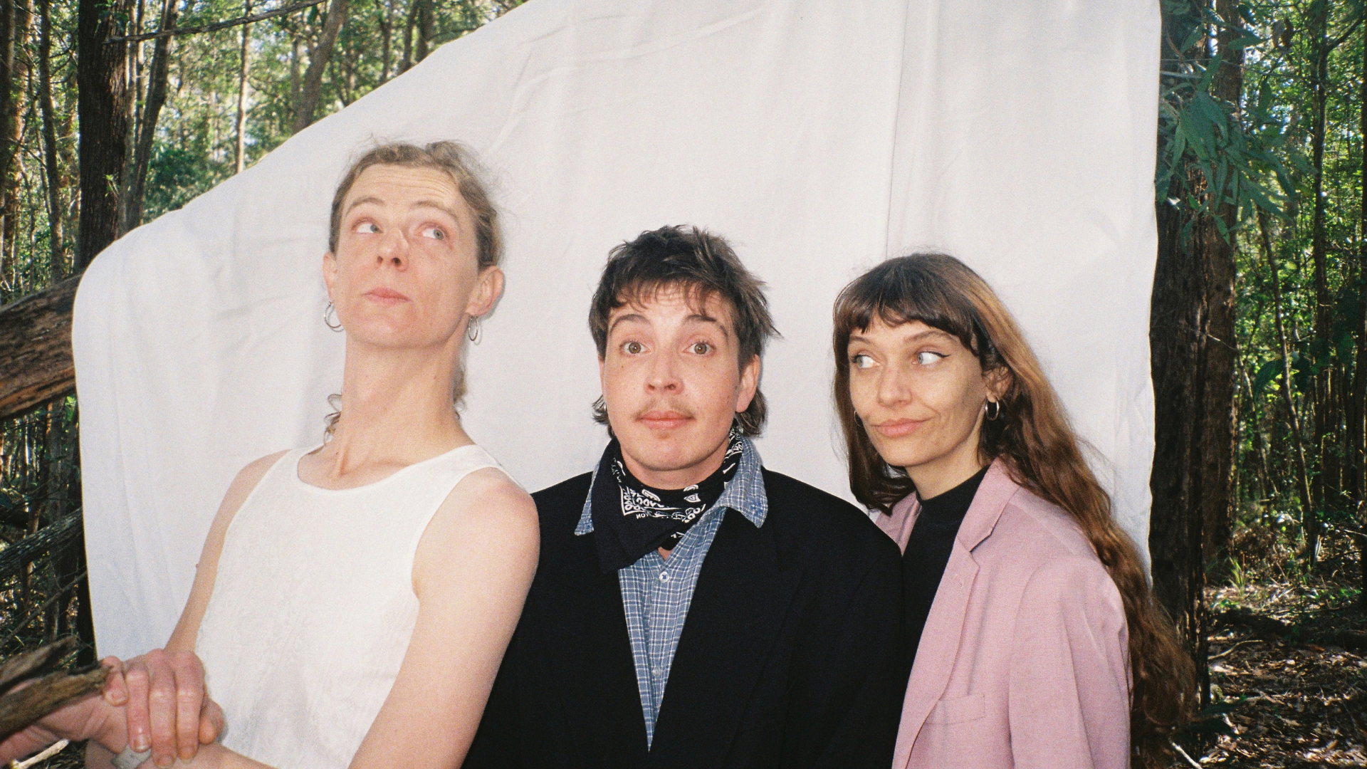 Three people stand in front of a white sheet hung in a forest. The person on the left wears a white sleeveless top, the middle person is in a suit with a scarf, and the right person wears a pink blazer. All look in different directions.