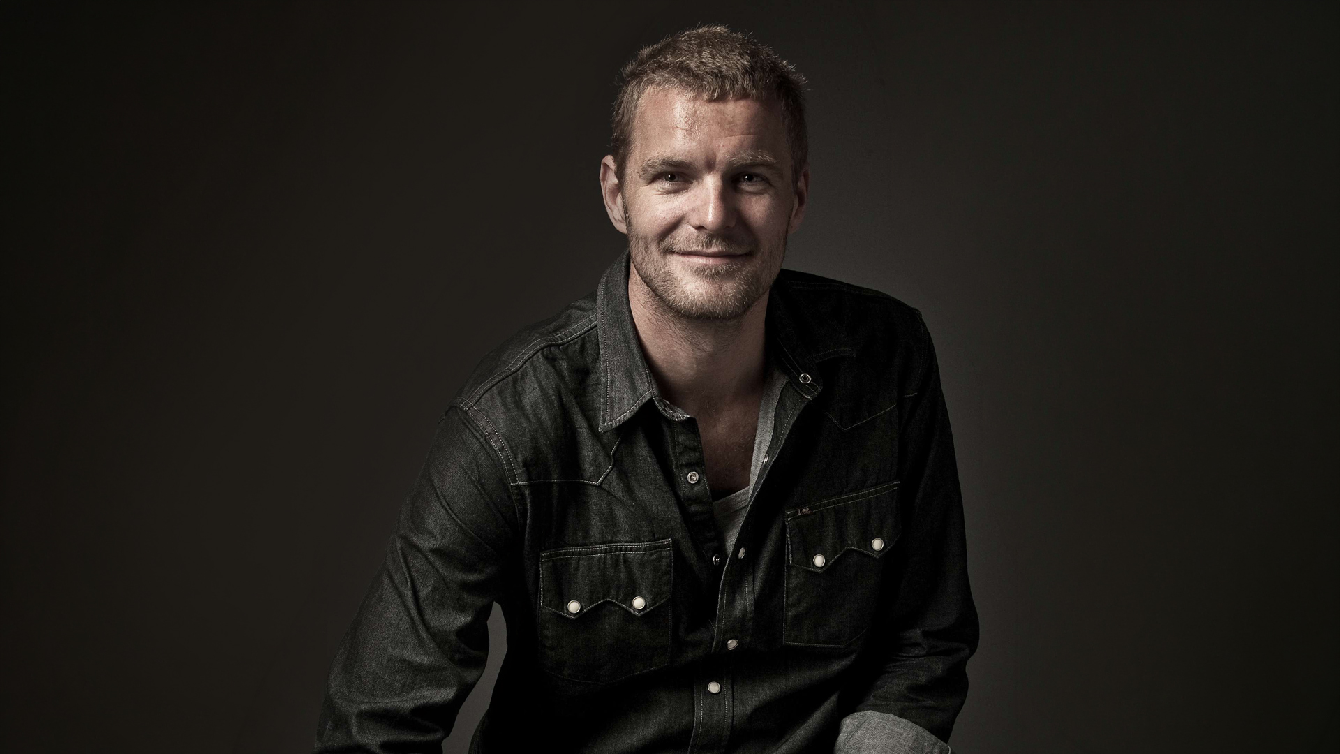 A man wearing a dark denim button-down shirt smiles at the camera against a dark background.